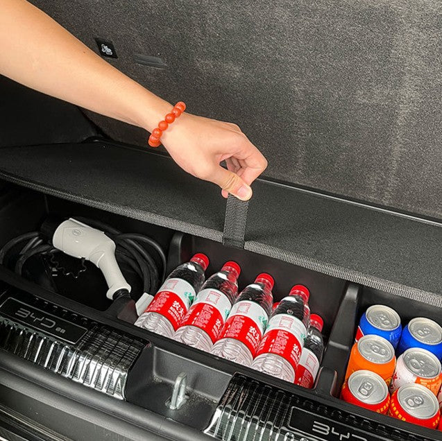 Hand pulling the lift strap on a BYD ATTO 1 custom-fit trunk boot organizer, showing easy access to neatly stored charging cable and drinks for tidy cargo storage.